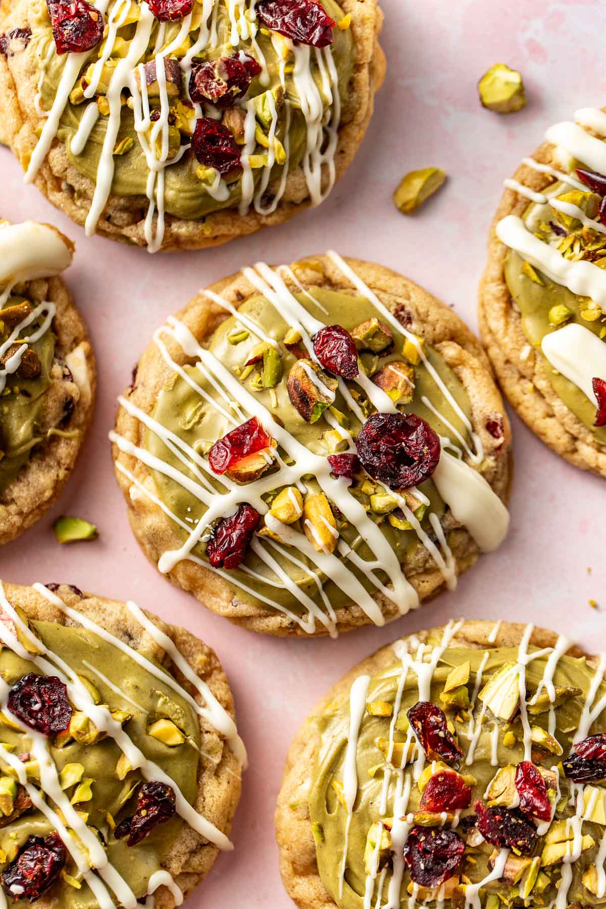 Close-up of pistachio cranberry cookies with white chocolate drizzle and bright red cranberries, surrounded by scattered pistachio pieces.