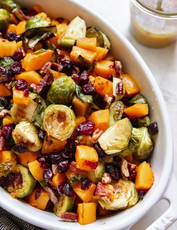 roasted brussels sprouts and butternut squash in a white baking dish garnished with chopped pecans and dried cranberries
