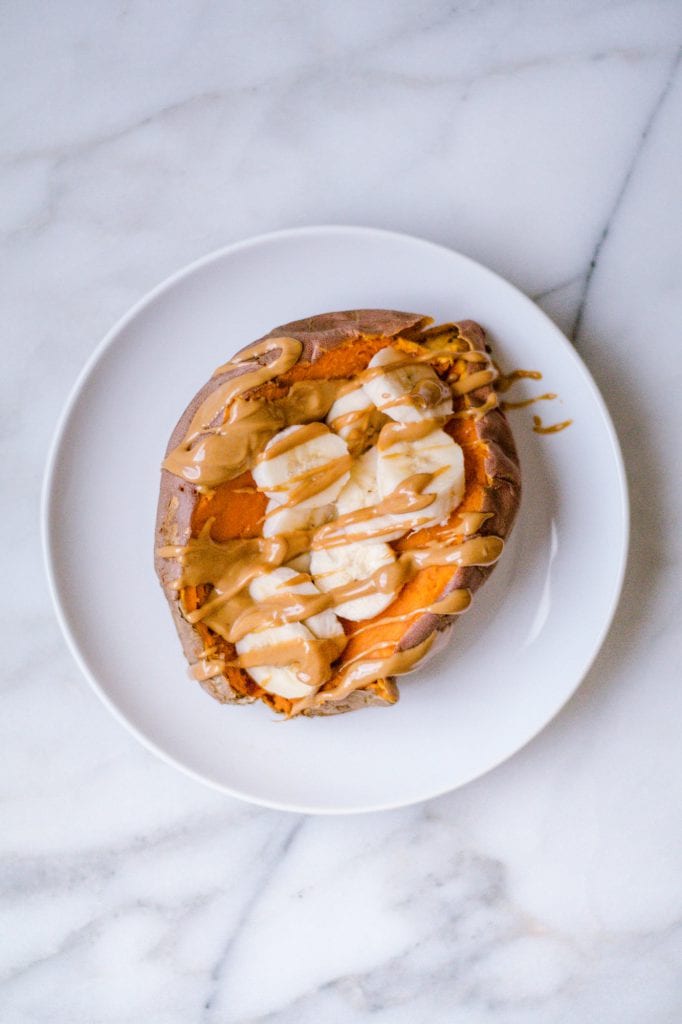 Cooked sweet potato sliced in half with banana slices on top and peanut butter drizzled over the banana. On a white plate. 