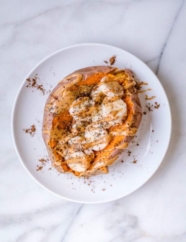 A sweet potato on a white plate filled with banana slices, peanut butter, chia seeds and cinnamon.