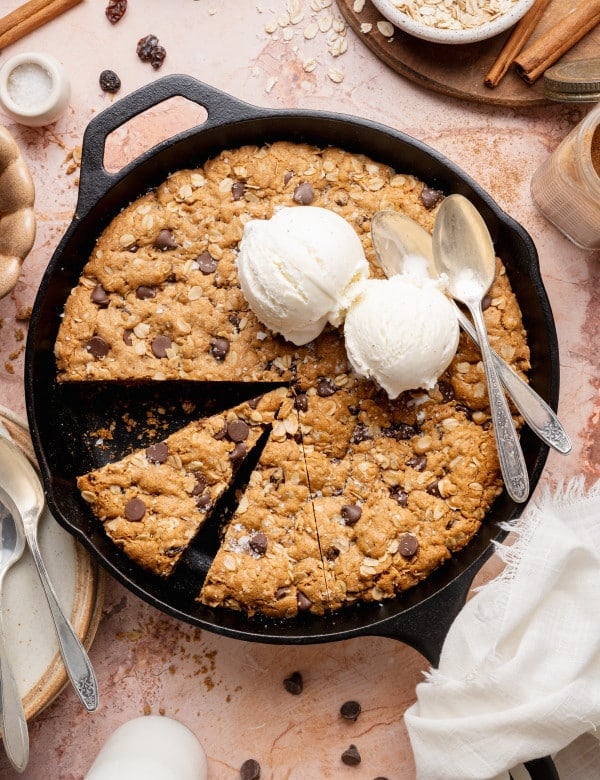An overhead view of an oatmeal raisin chocolate chip cookie skillet baked in a black cast iron pan, with a slice cut out and two scoops of vanilla ice cream melting on top, surrounded by cinnamon sticks, oats, chocolate chips, and baking props on a neutral surface.
