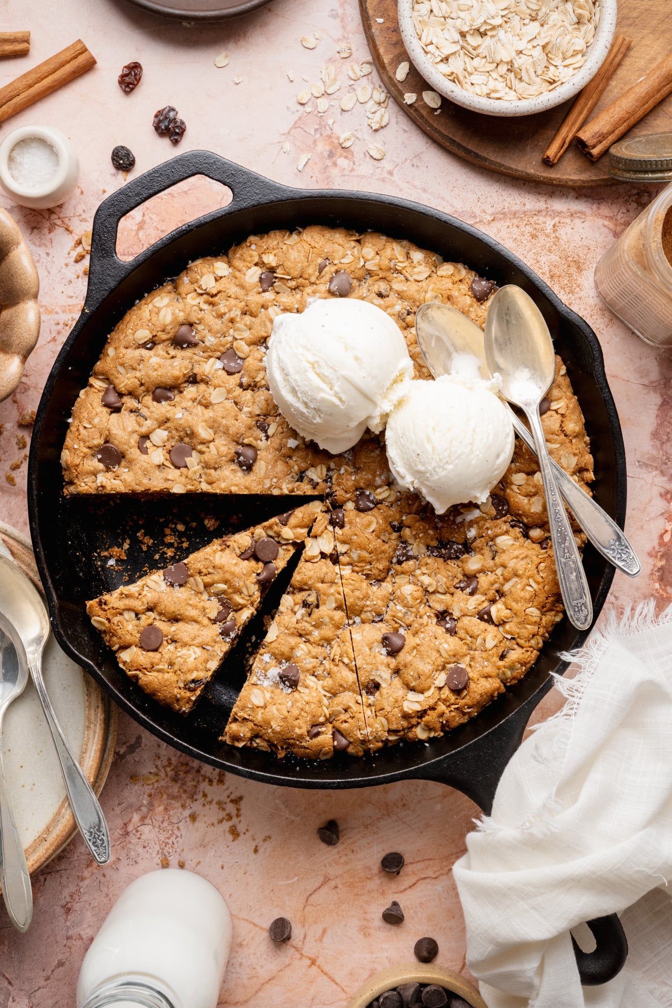 An overhead view of an oatmeal raisin chocolate chip cookie skillet baked in a black cast iron pan, with a slice cut out and two scoops of vanilla ice cream melting on top, surrounded by cinnamon sticks, oats, chocolate chips, and baking props on a neutral surface.