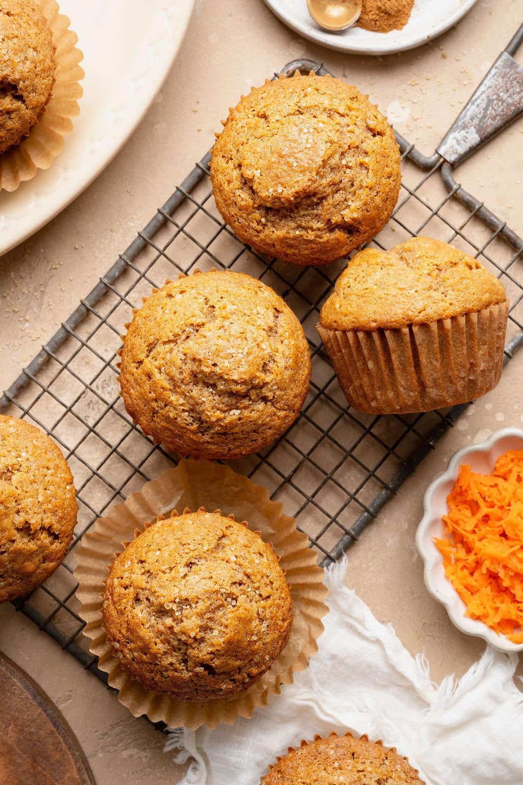 Overhead view of carrot cake muffins cooling on a wire rack, topped with coarse sugar, with grated carrots and cinnamon nearby on a neutral background.