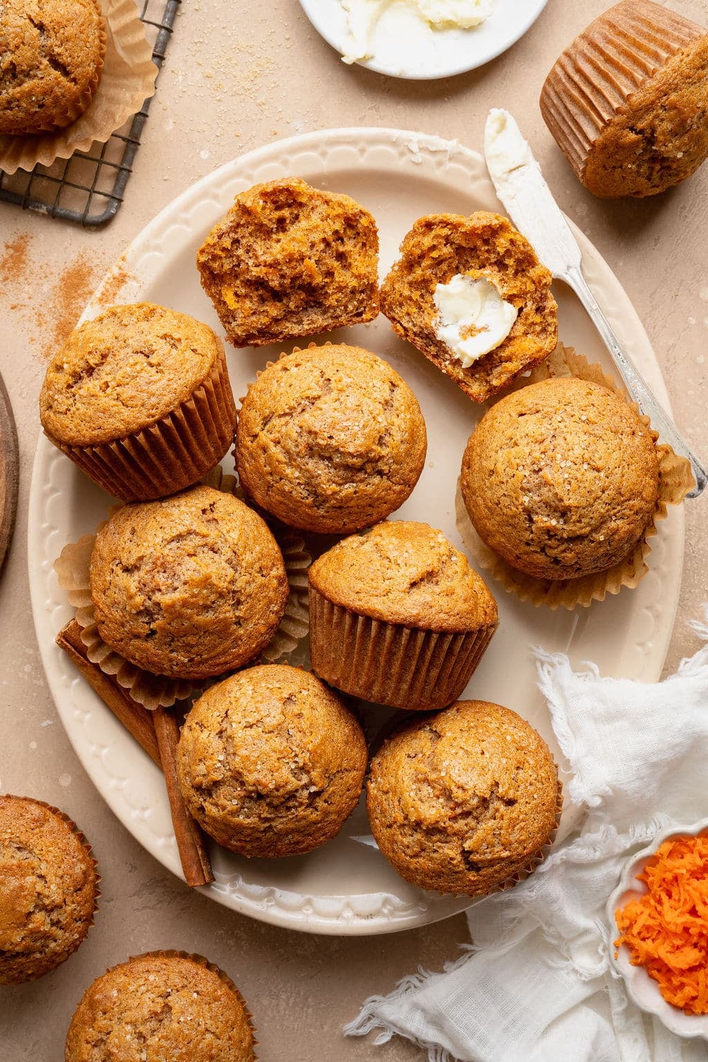 Platter of carrot cake muffins with one split open and spread with butter, surrounded by cinnamon sticks, grated carrots, and a butter knife.
