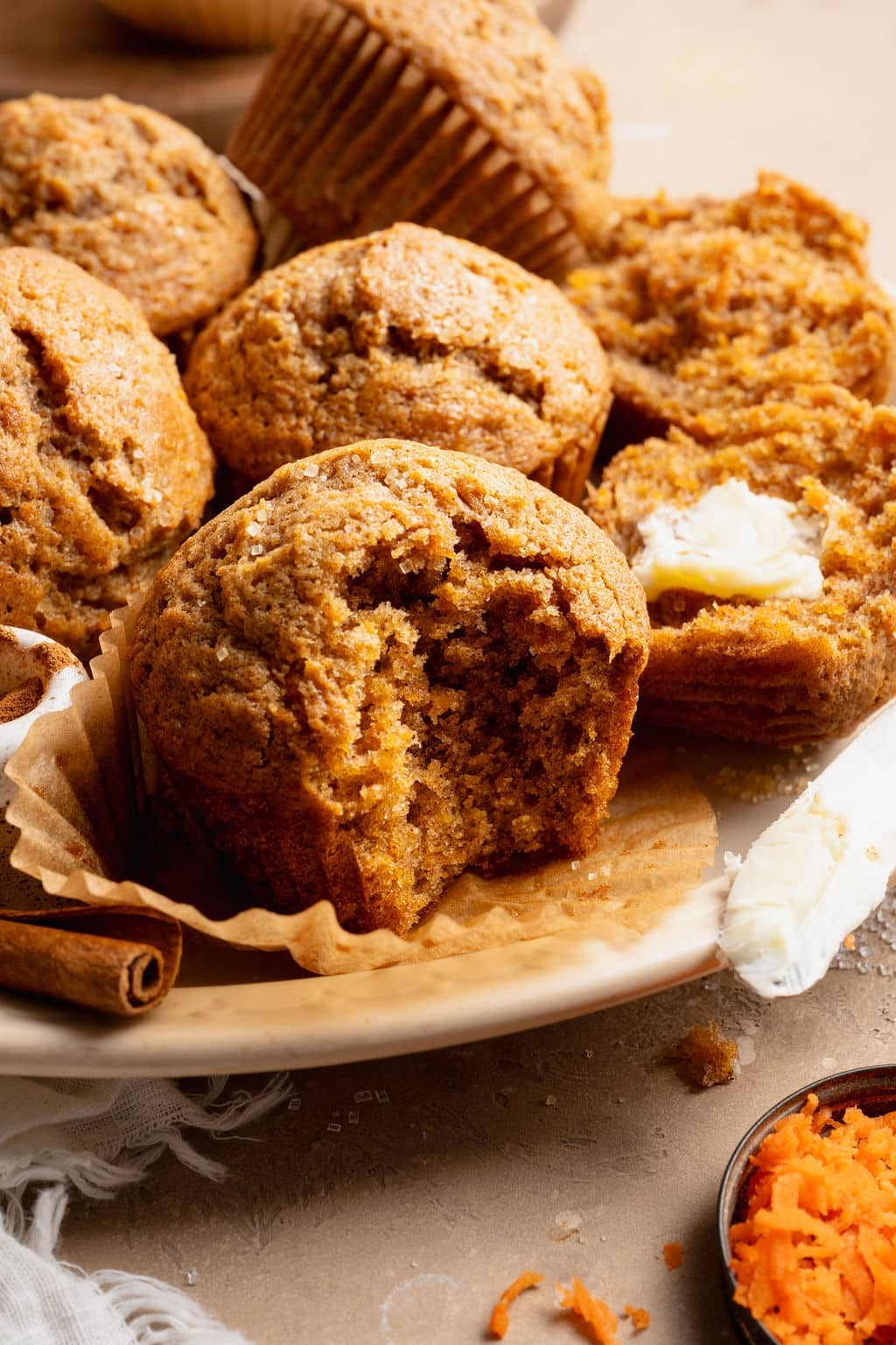 Close-up of a carrot cake muffin with a bite taken out, showing a moist, tender crumb, served on a plate with butter and cinnamon sticks.