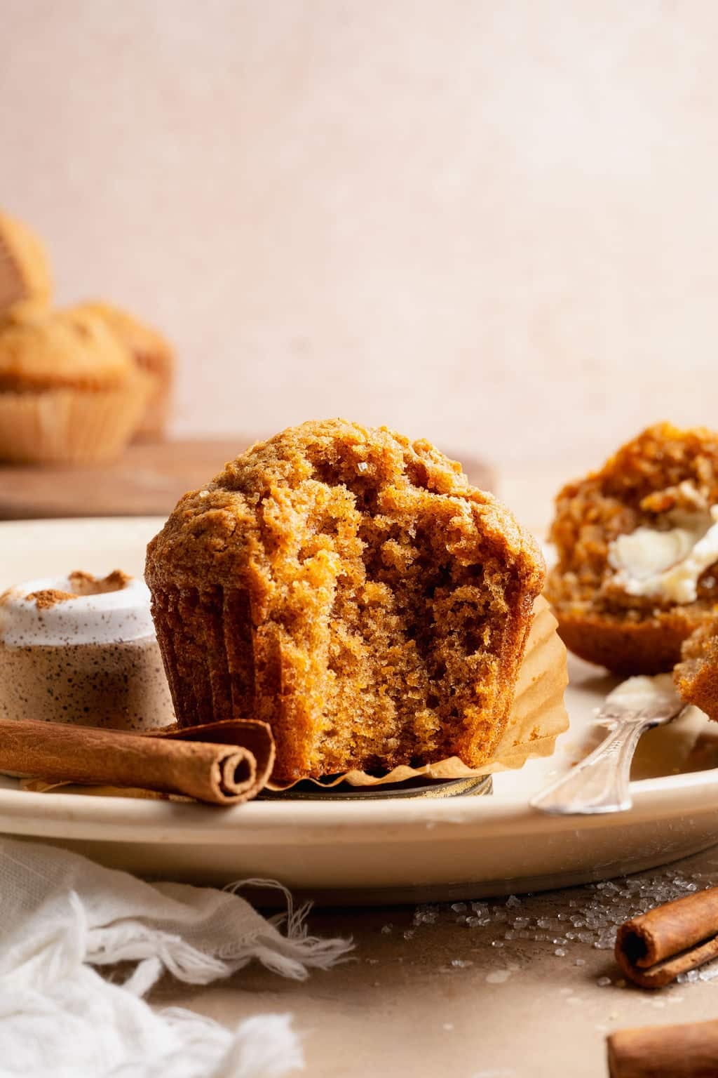 Single carrot cake muffin on a plate with cinnamon sticks and a small jar of cinnamon in the background, styled with soft neutral linens.