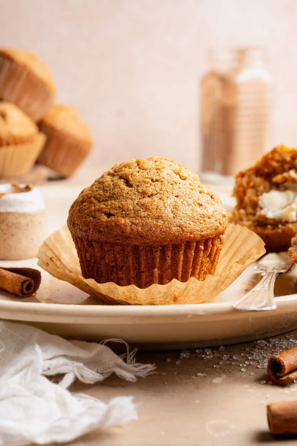 Side view of a carrot cake muffin in a paper liner on a plate, with another muffin split open and buttered in the background.