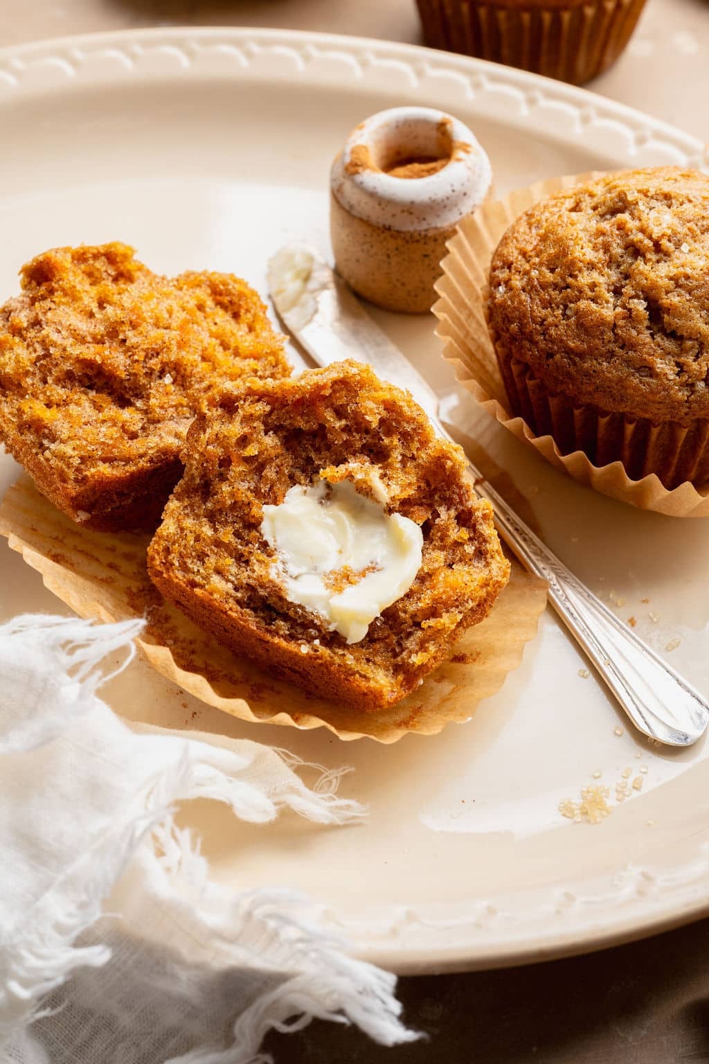 Carrot cake muffin broken in half and topped with softened butter on a parchment liner, with a butter knife and cinnamon jar nearby.