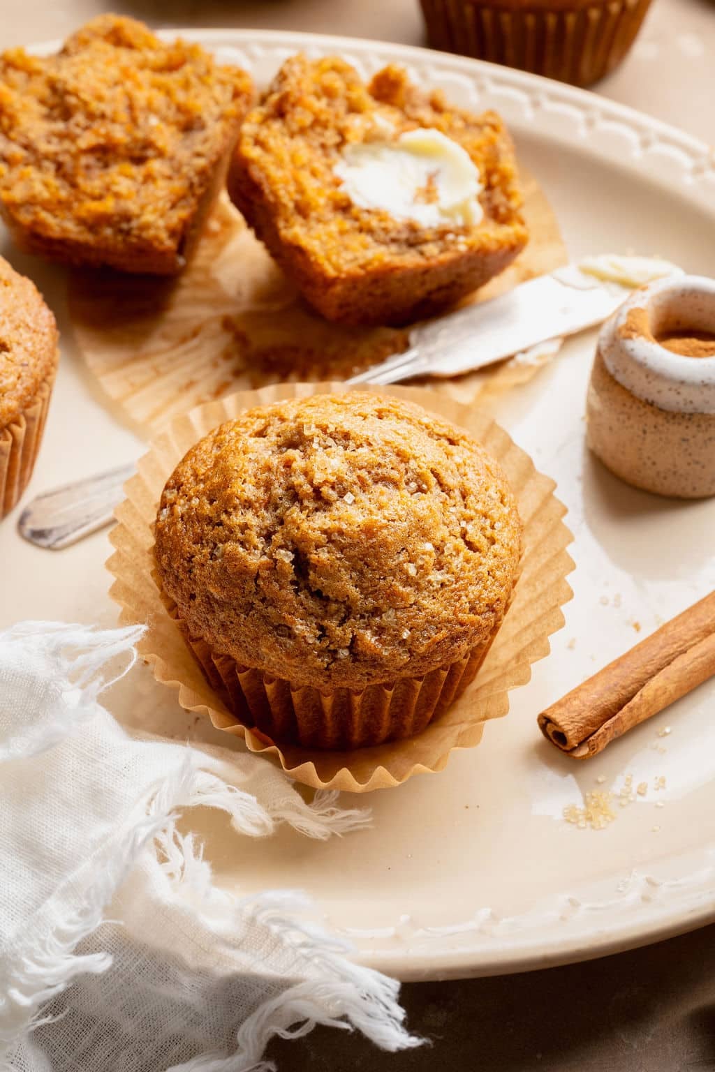 Single carrot cake muffin on a plate with cinnamon sticks and a split muffin in the background, highlighting the domed top and sugar crust.