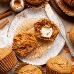 Single carrot cake muffin on a plate with cinnamon sticks and a split muffin in the background, highlighting the domed top and sugar crust.