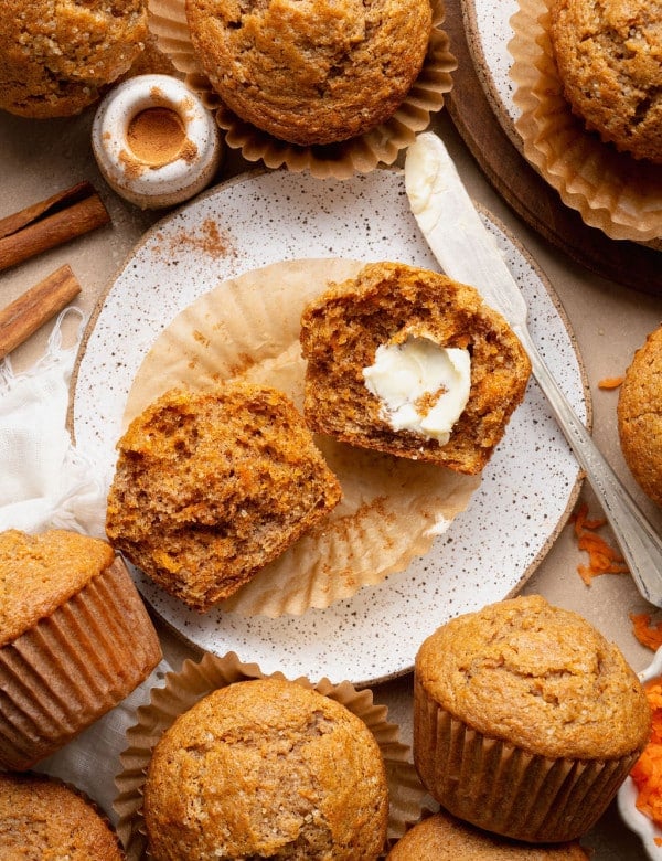 Single carrot cake muffin on a plate with cinnamon sticks and a split muffin in the background, highlighting the domed top and sugar crust.