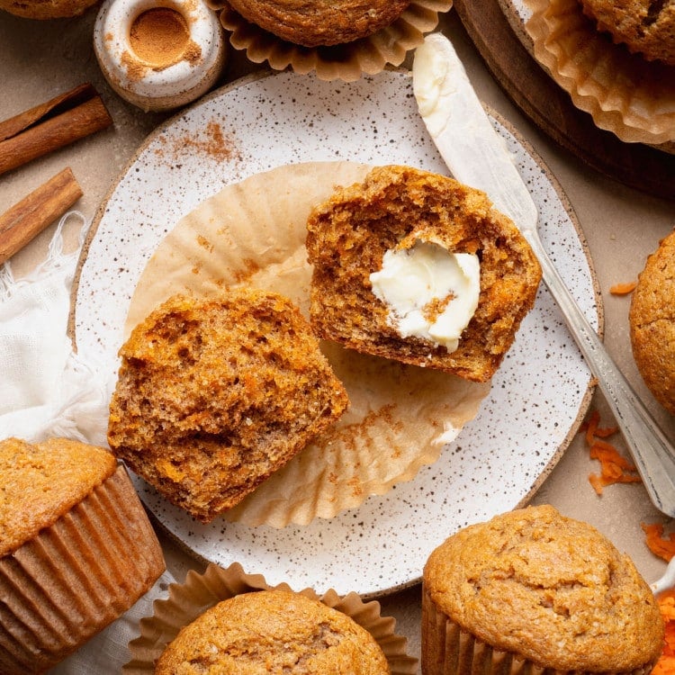 Single carrot cake muffin on a plate with cinnamon sticks and a split muffin in the background, highlighting the domed top and sugar crust.