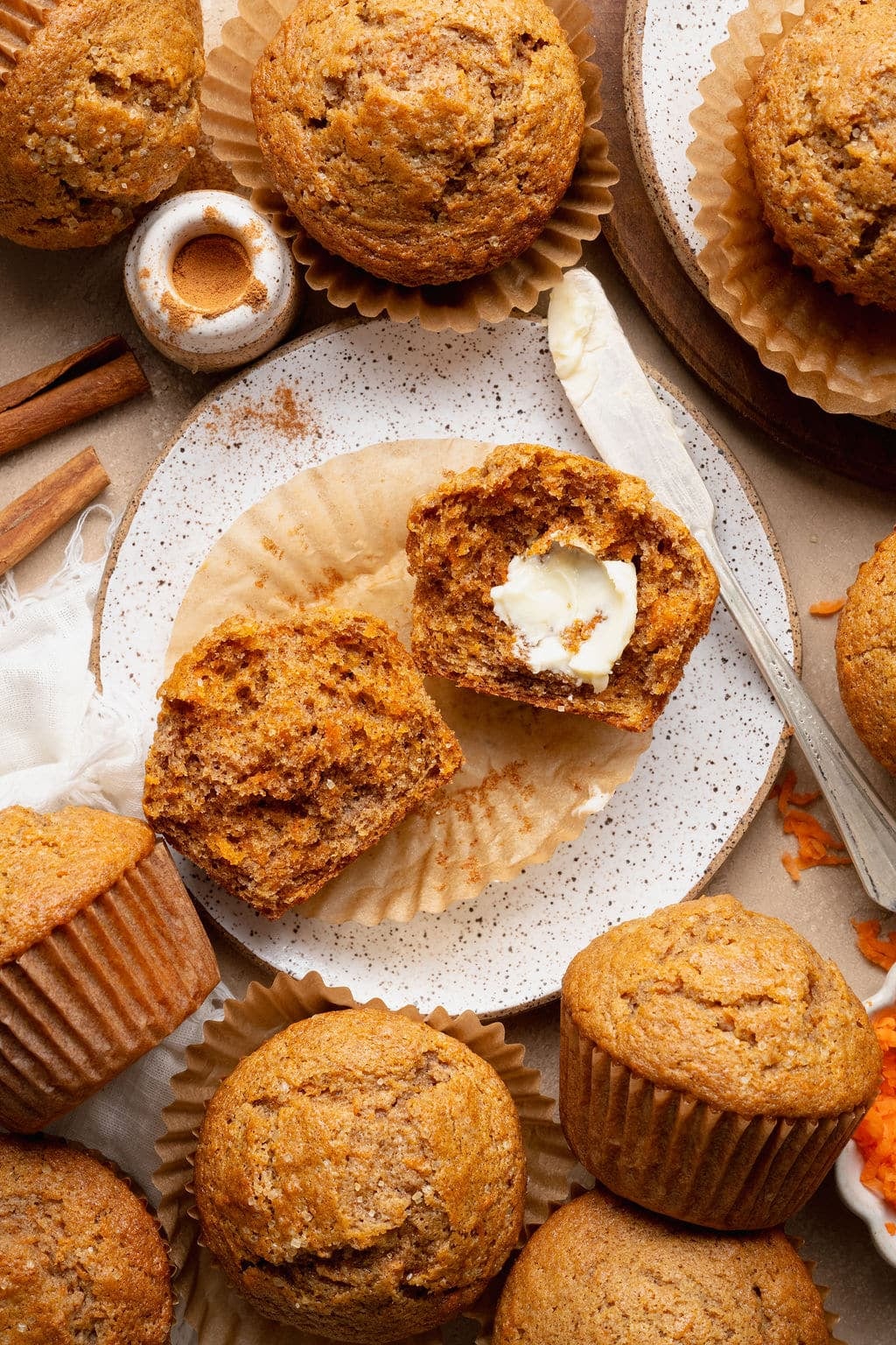 Single carrot cake muffin on a plate with cinnamon sticks and a split muffin in the background, highlighting the domed top and sugar crust.