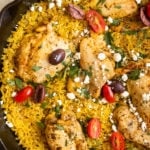 chicken thighs and yellow rice in a cast iron skillet garnished with feta, parsley, tomatoes and kalamata olives