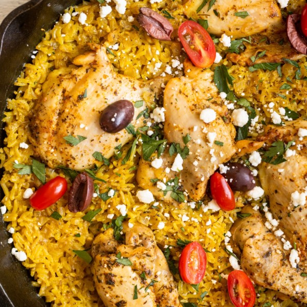 chicken thighs and yellow rice in a cast iron skillet garnished with feta, parsley, tomatoes and kalamata olives