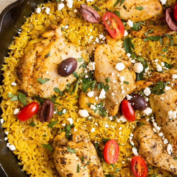 chicken thighs and yellow rice in a cast iron skillet garnished with feta, parsley, tomatoes and kalamata olives