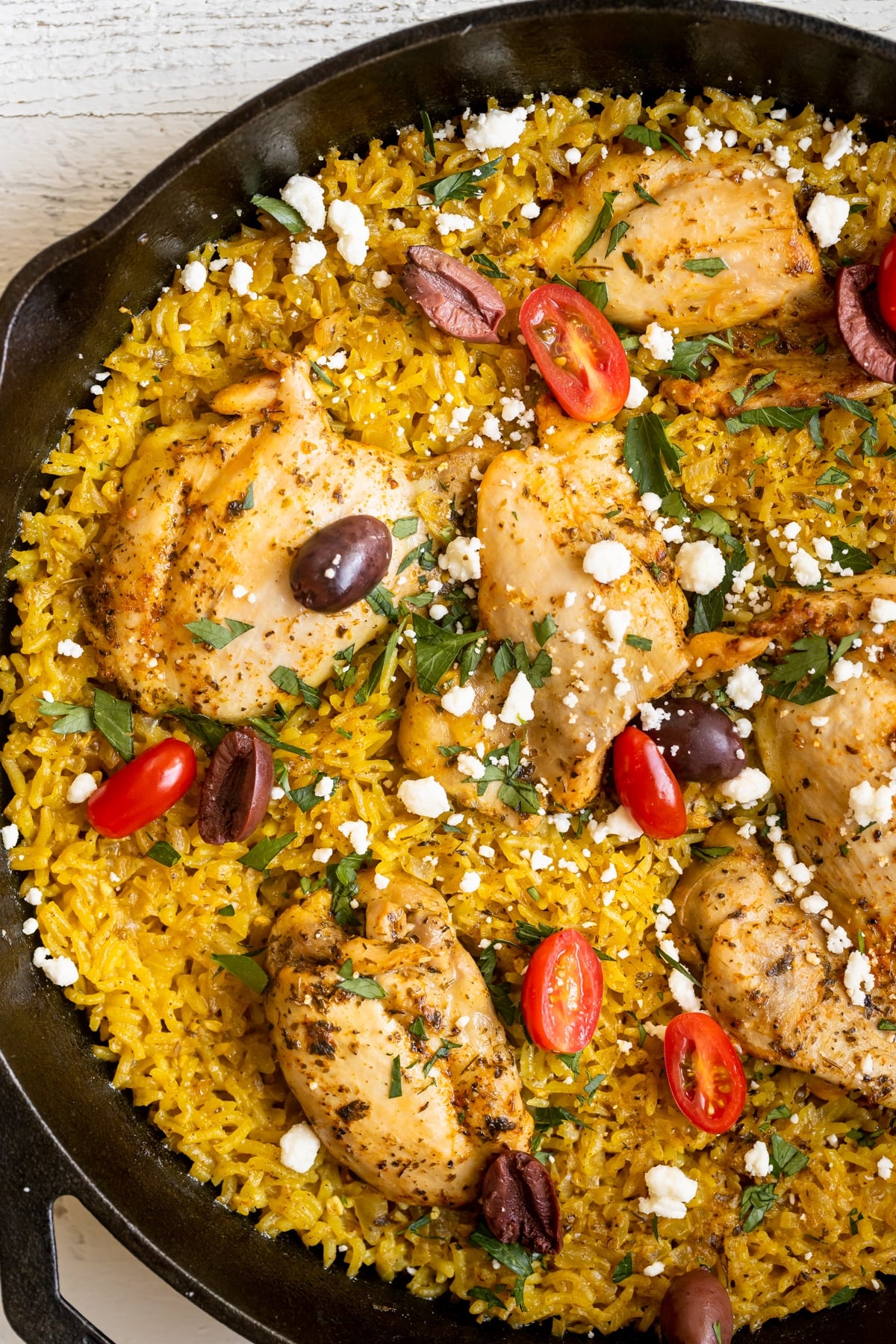 chicken thighs and yellow rice in a cast iron skillet garnished with feta, parsley, tomatoes and kalamata olives