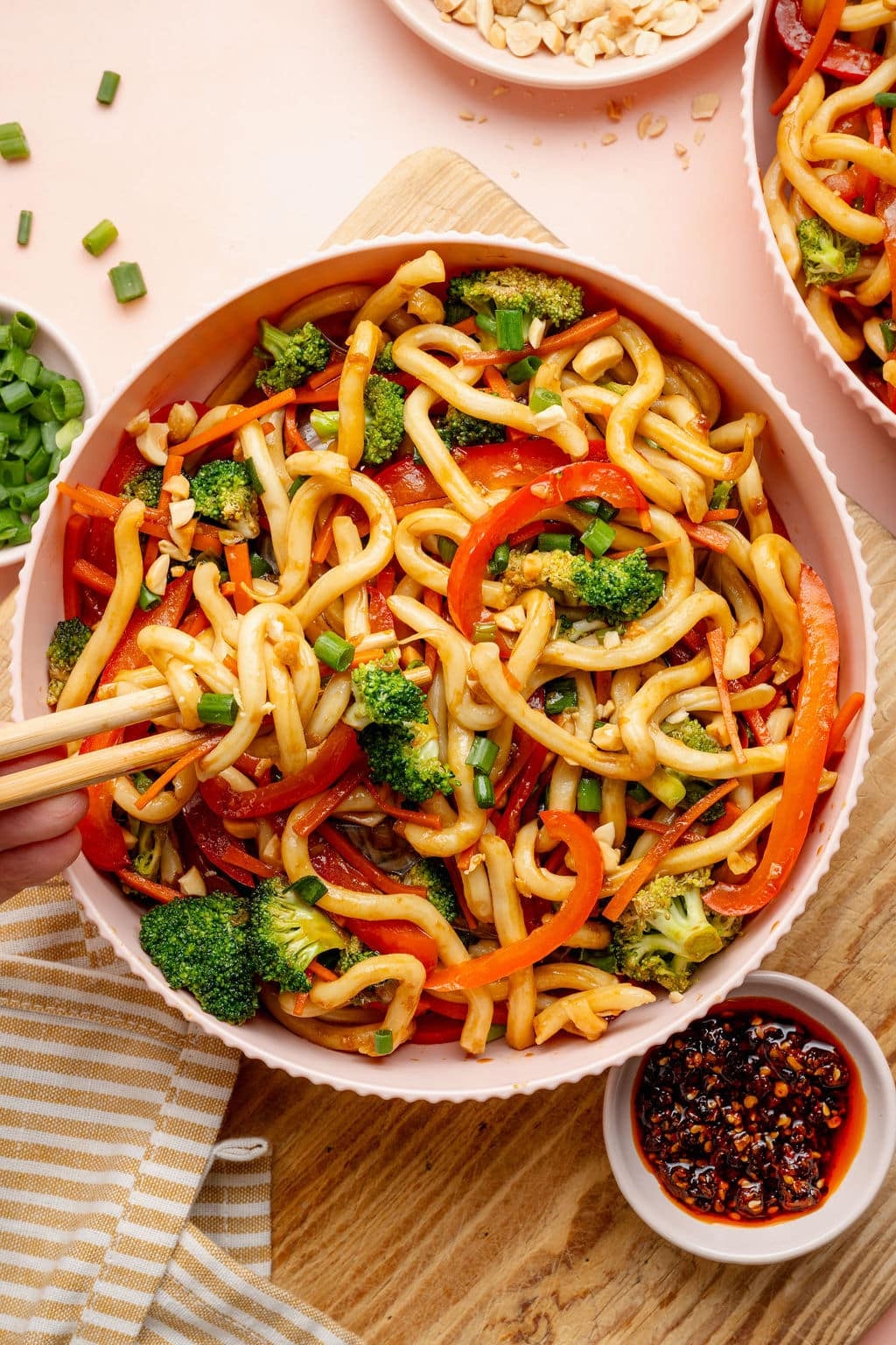 Overhead image of a bowl of sesame udon noodles with broccoli, carrots, and red bell peppers. Chopsticks lift a portion of noodles from the bowl, with chili crisp and chopped peanuts nearby.