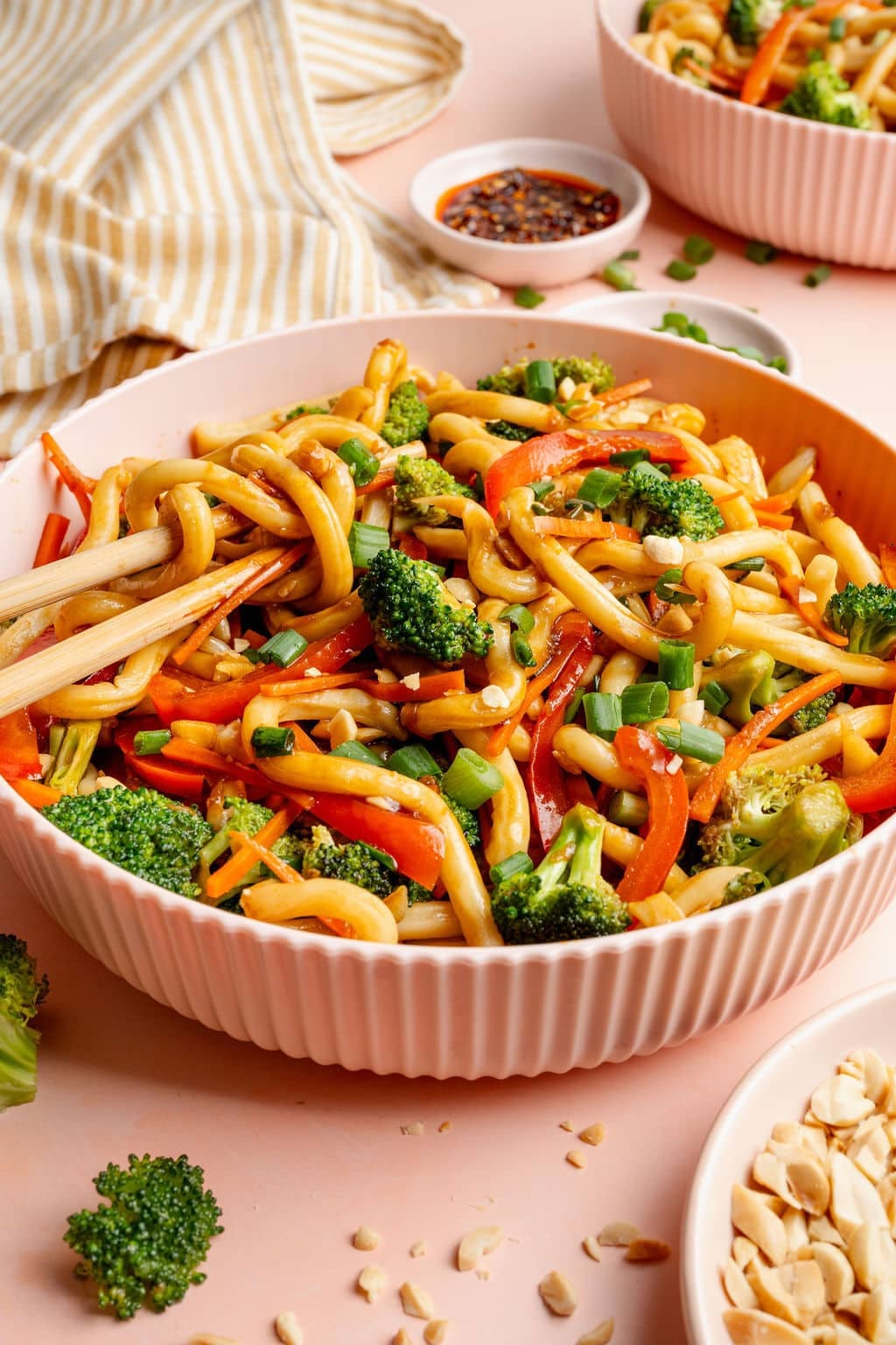 Side-angle image of a bowl of sesame udon noodles with broccoli, carrots, and red bell peppers. Chopsticks lift noodles, with a striped kitchen towel and bowls of chili crisp and chopped peanuts in the background.