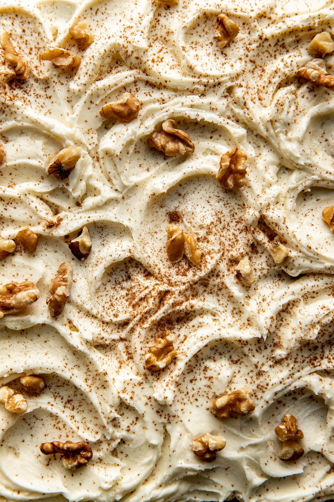Close-up of creamy cream cheese frosting swirled over carrot cake bars, topped with walnut pieces and a dusting of cinnamon.
