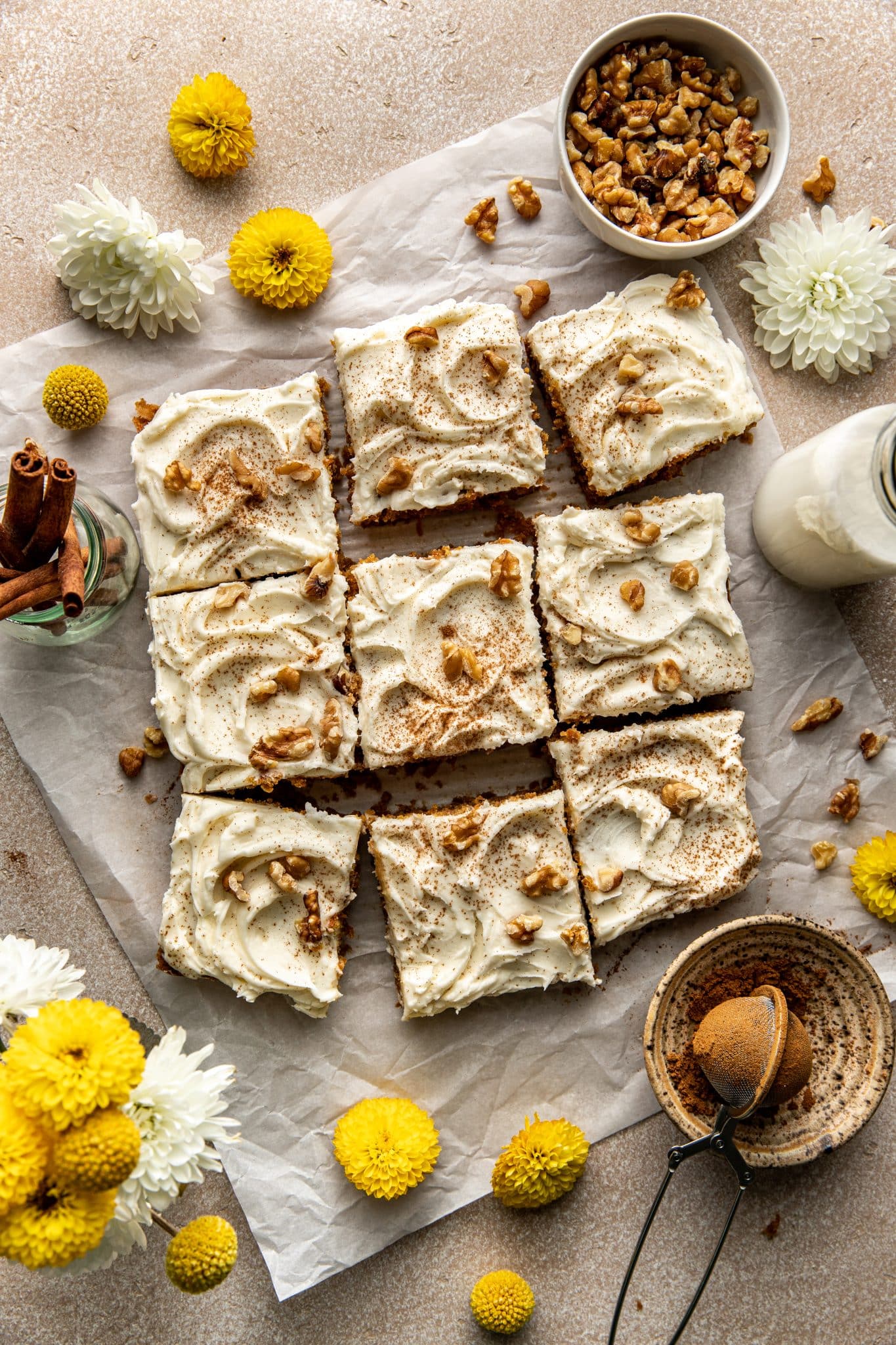 Carrot cake bars cut into squares on parchment paper, topped with cream cheese frosting, cinnamon, and chopped walnuts, surrounded by fresh flowers and cinnamon sticks.