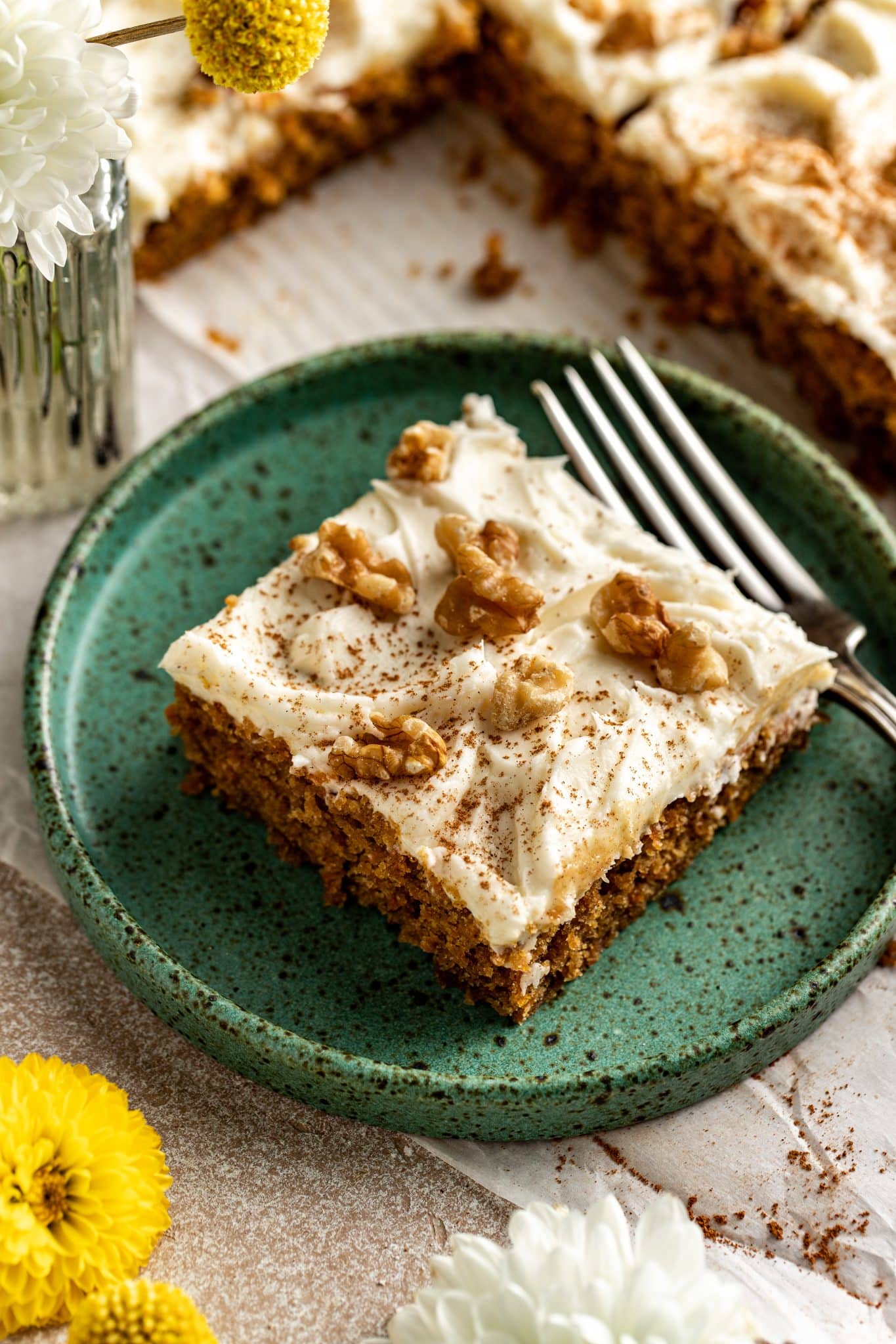 Single slice of carrot cake bar on a rustic green plate with a fork, topped with frosting, walnuts, and cinnamon, with flowers in the background.