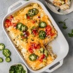 white dish with a mexican casserole inside & a piece missing, topped with avocado, tomatoes, and shredded cheese