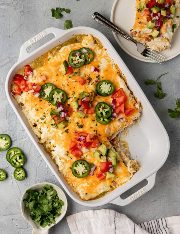 white dish with a mexican casserole inside & a piece missing, topped with avocado, tomatoes, and shredded cheese