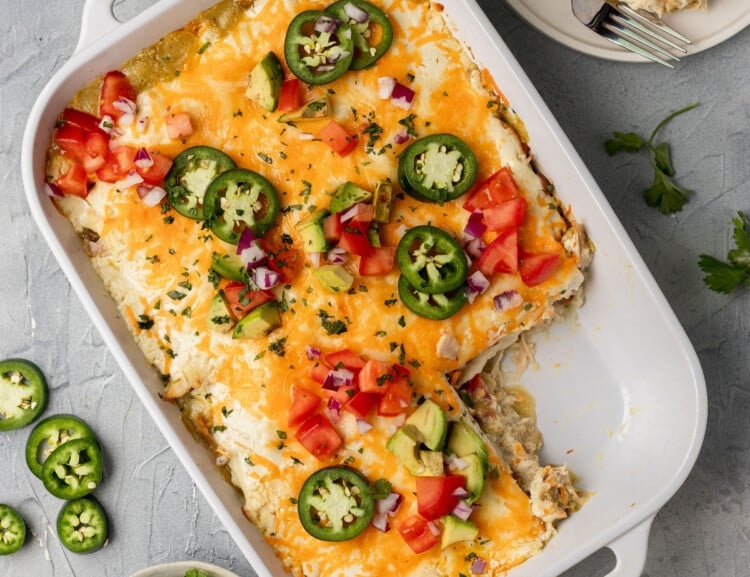 white dish with a mexican casserole inside & a piece missing, topped with avocado, tomatoes, and shredded cheese