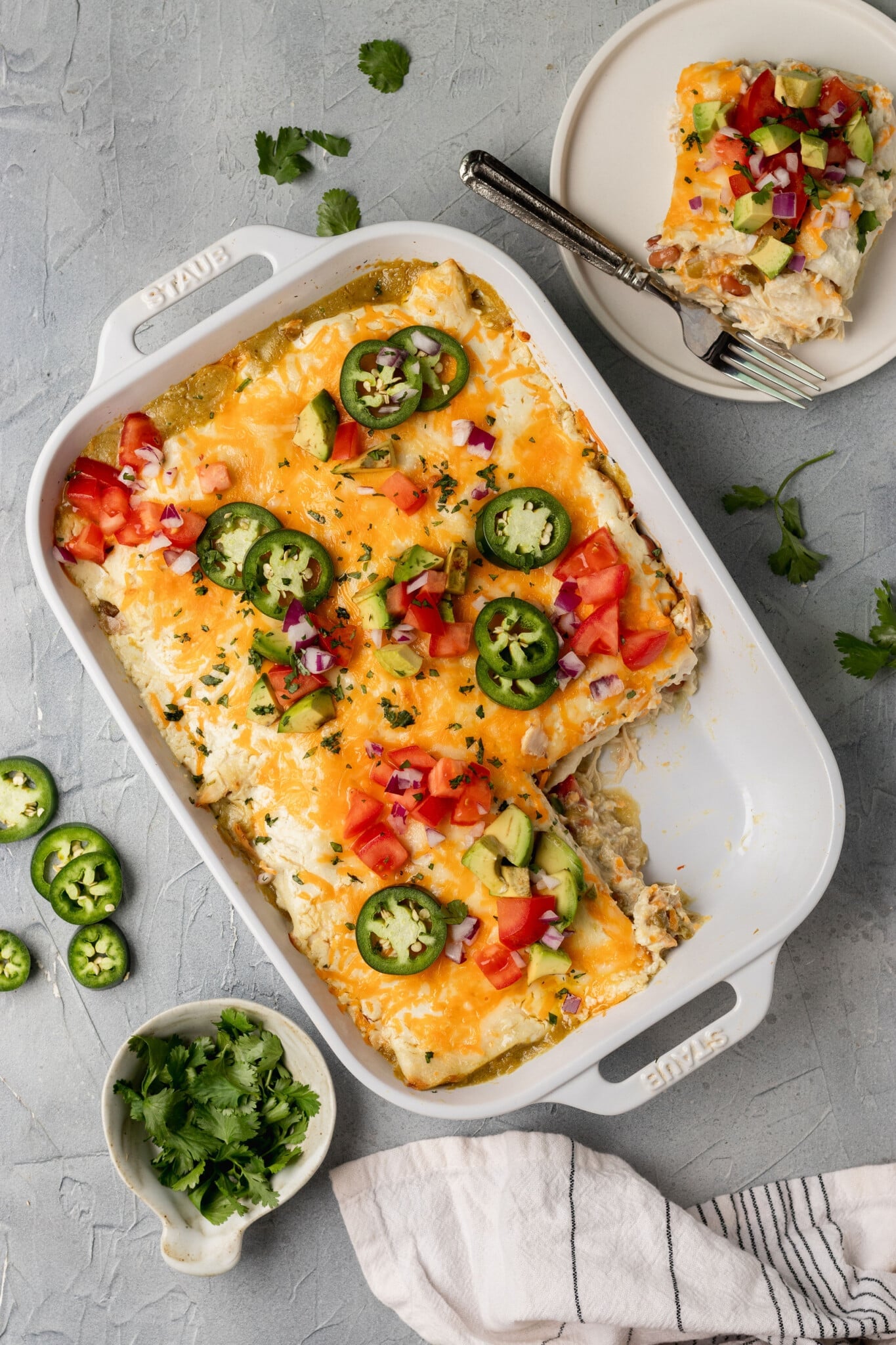 white dish with a mexican casserole inside & a piece missing, topped with avocado, tomatoes, and shredded cheese