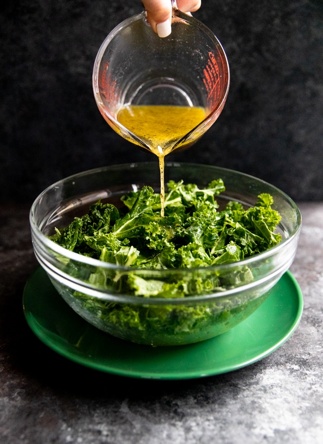 massaged kale in a glass bowl on a green plate with lemon poppyseed dressing being poured on top