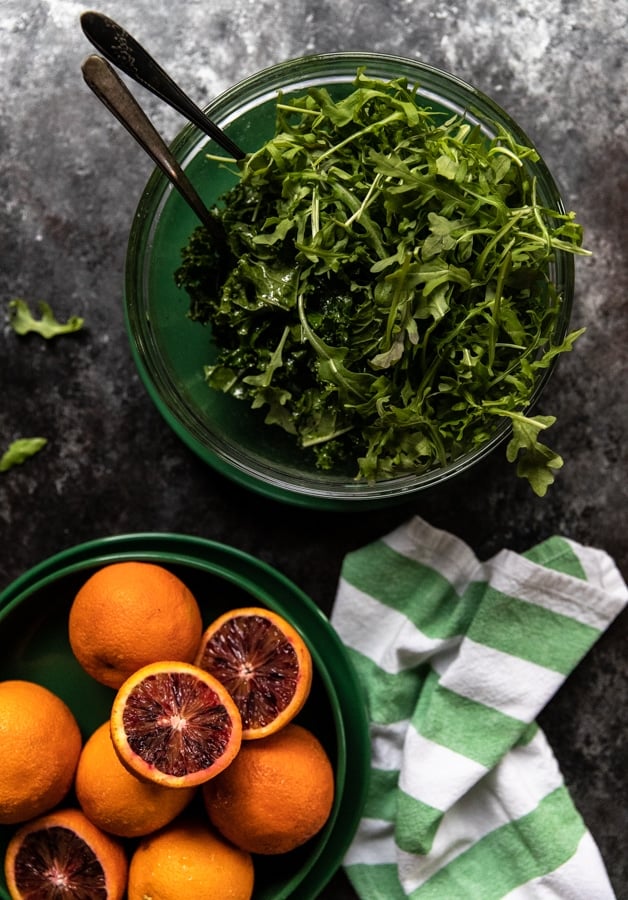 arugula and kale in a glass bowl on top of a green plate with a side of blood oranges