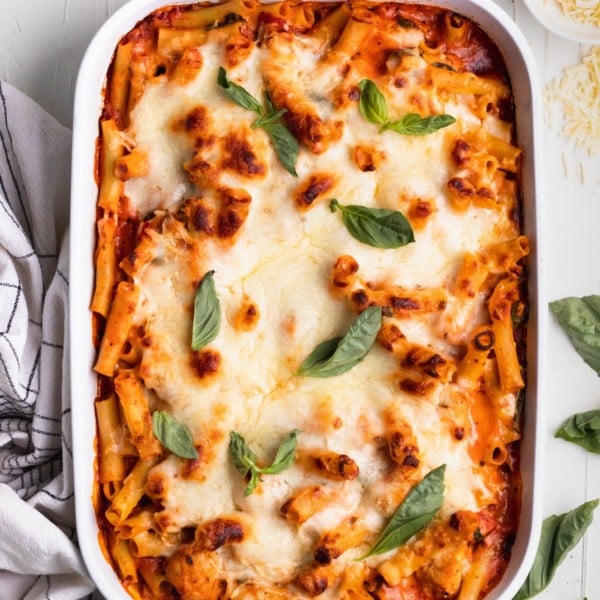 baked ziti in a white casserole dish topped with cheese and fresh basil