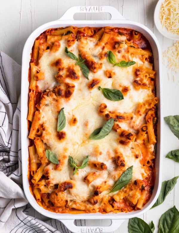 baked ziti in a white casserole dish topped with cheese and fresh basil