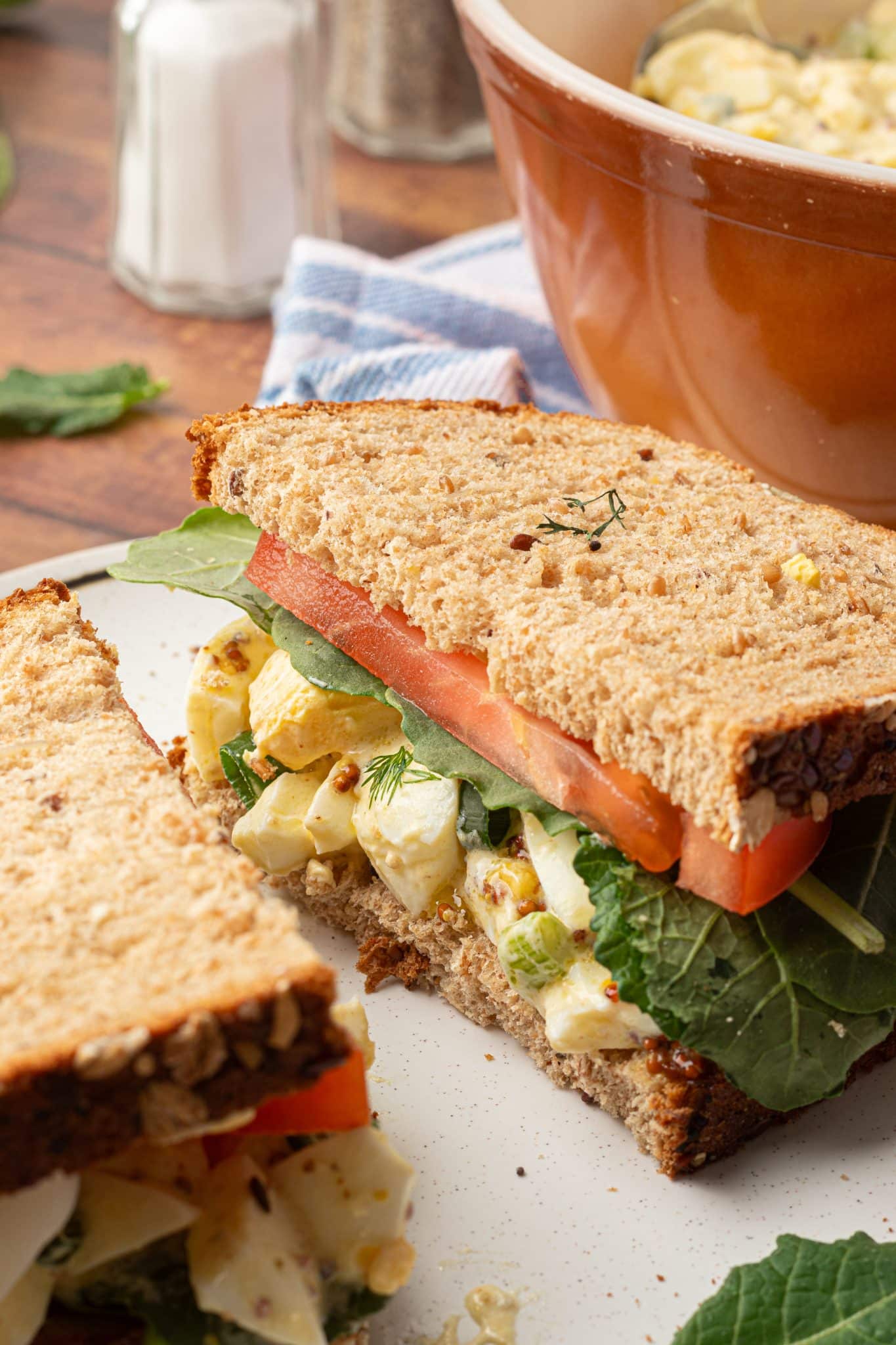 A close-up of a sliced egg salad sandwich on whole-grain bread, layered with leafy greens and fresh tomato slices, with a bowl of extra egg salad in the background. Salt and pepper shakers rest nearby on a wooden surface.







