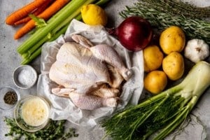 raw whole chicken on parchment paper surrounded by uncut veggies and spices