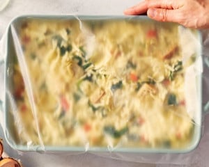 Overhead image of the assembled overnight artichoke spinach egg bake in a blue casserole dish covered tightly with plastic wrap before refrigerating overnight.