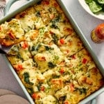 Overhead photo of a baked overnight artichoke spinach egg bake in a light blue casserole dish with a square slice lifted on a spatula. The cheesy breakfast casserole is filled with spinach, marinated artichokes, red bell peppers, toasted English muffin pieces, and chopped chives sprinkled on top.