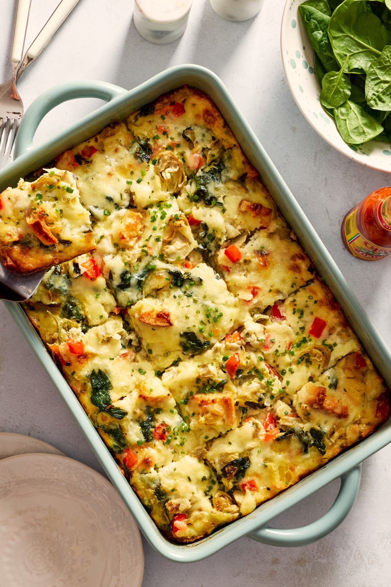 Overhead photo of a baked overnight artichoke spinach egg bake in a light blue casserole dish with a square slice lifted on a spatula. The cheesy breakfast casserole is filled with spinach, marinated artichokes, red bell peppers, toasted English muffin pieces, and chopped chives sprinkled on top.