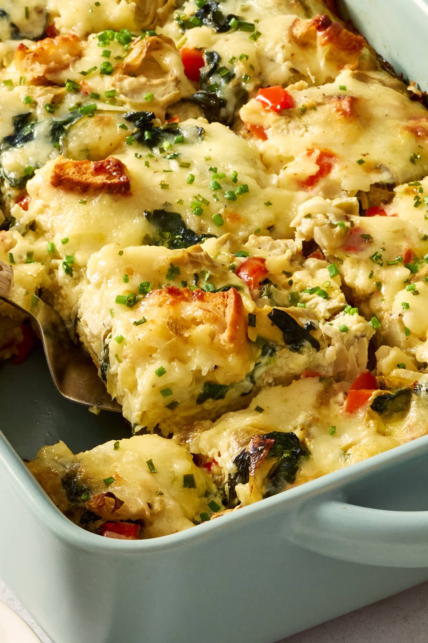 Close-up photo of a cheesy overnight artichoke spinach egg bake in a light blue casserole dish with a serving spoon lifting out a square portion. The casserole is filled with spinach, marinated artichokes, toasted English muffin pieces, red bell peppers, and chopped chives.