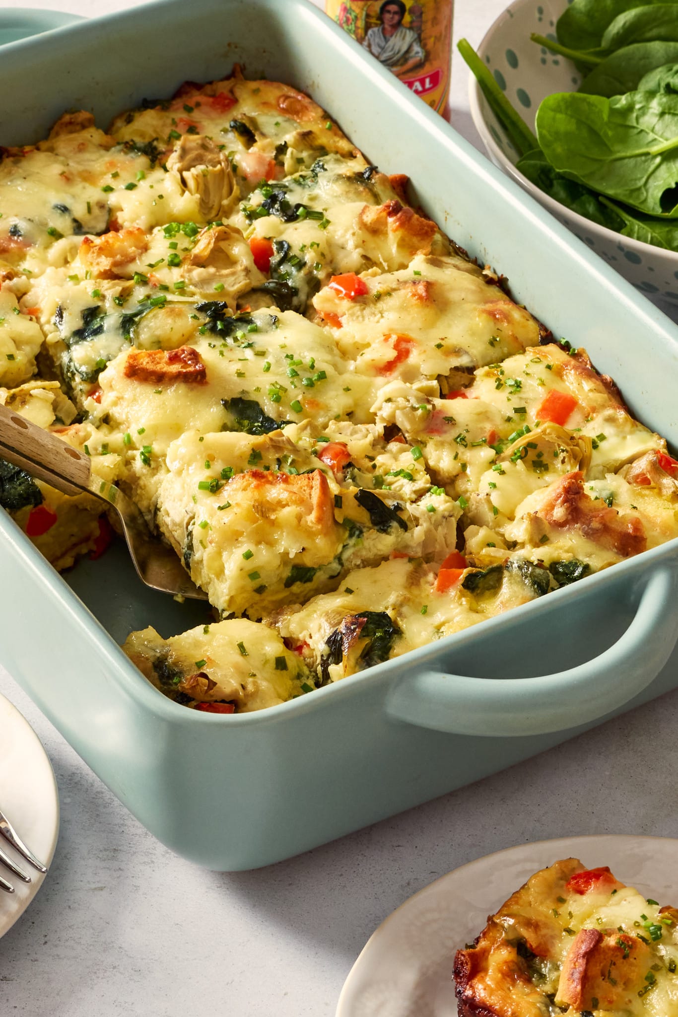 Wide overhead photo of a baked overnight artichoke spinach egg bake in a light blue casserole dish with a serving spoon scooping out a portion. A plated slice sits nearby alongside fresh spinach and a bottle of hot sauce.