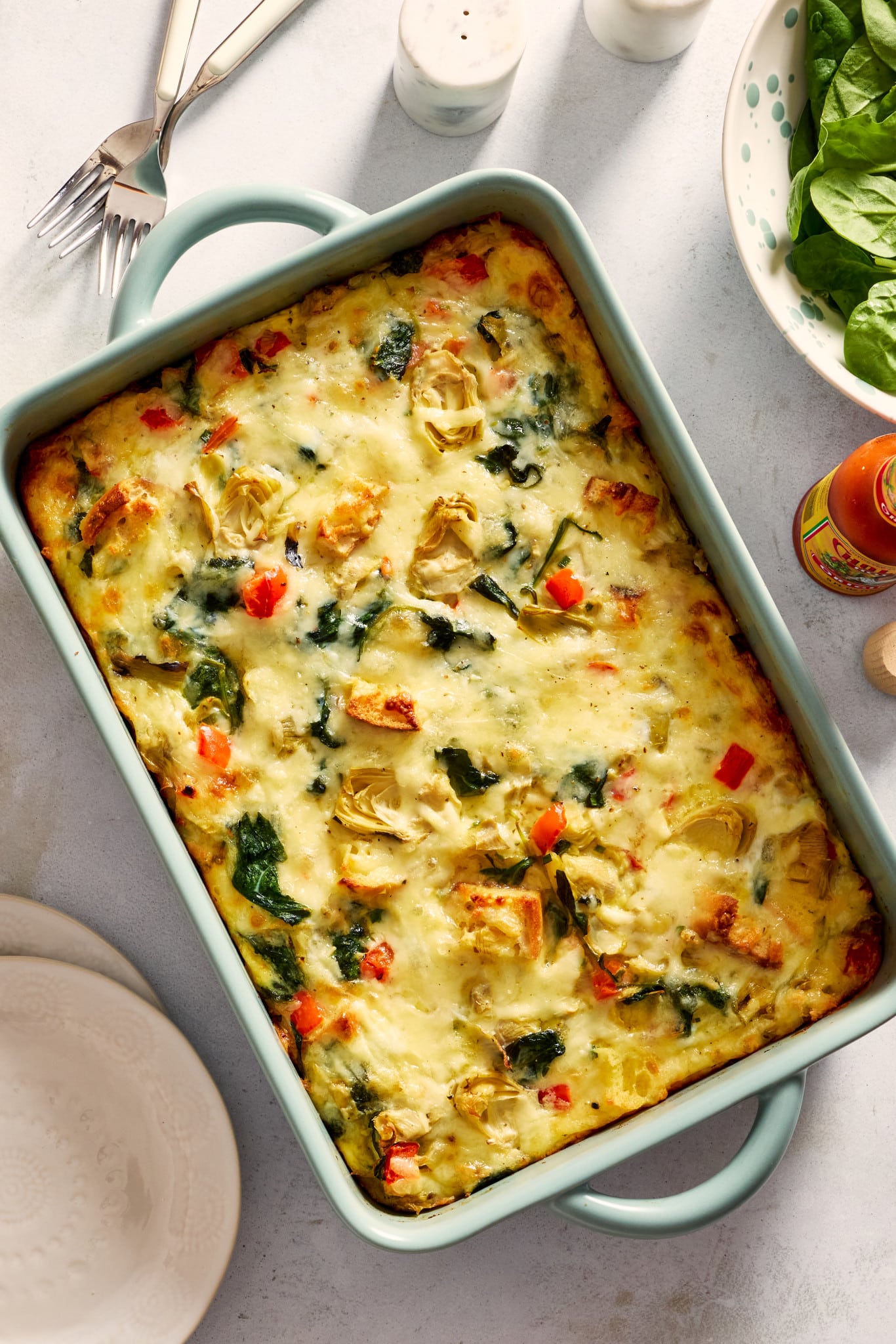 Overhead photo of a baked overnight artichoke spinach egg bake in a light blue casserole dish. The casserole is filled with melted cheese, spinach, marinated artichokes, toasted English muffin pieces, and diced red bell peppers.