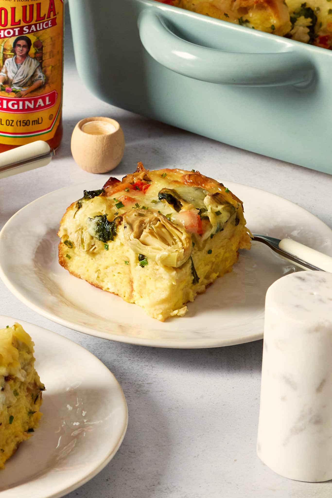 Close-up photo of a plated slice of overnight artichoke spinach egg bake topped with melted cheese, spinach, marinated artichokes, and red bell peppers. The casserole dish and bottle of hot sauce are visible in the background.