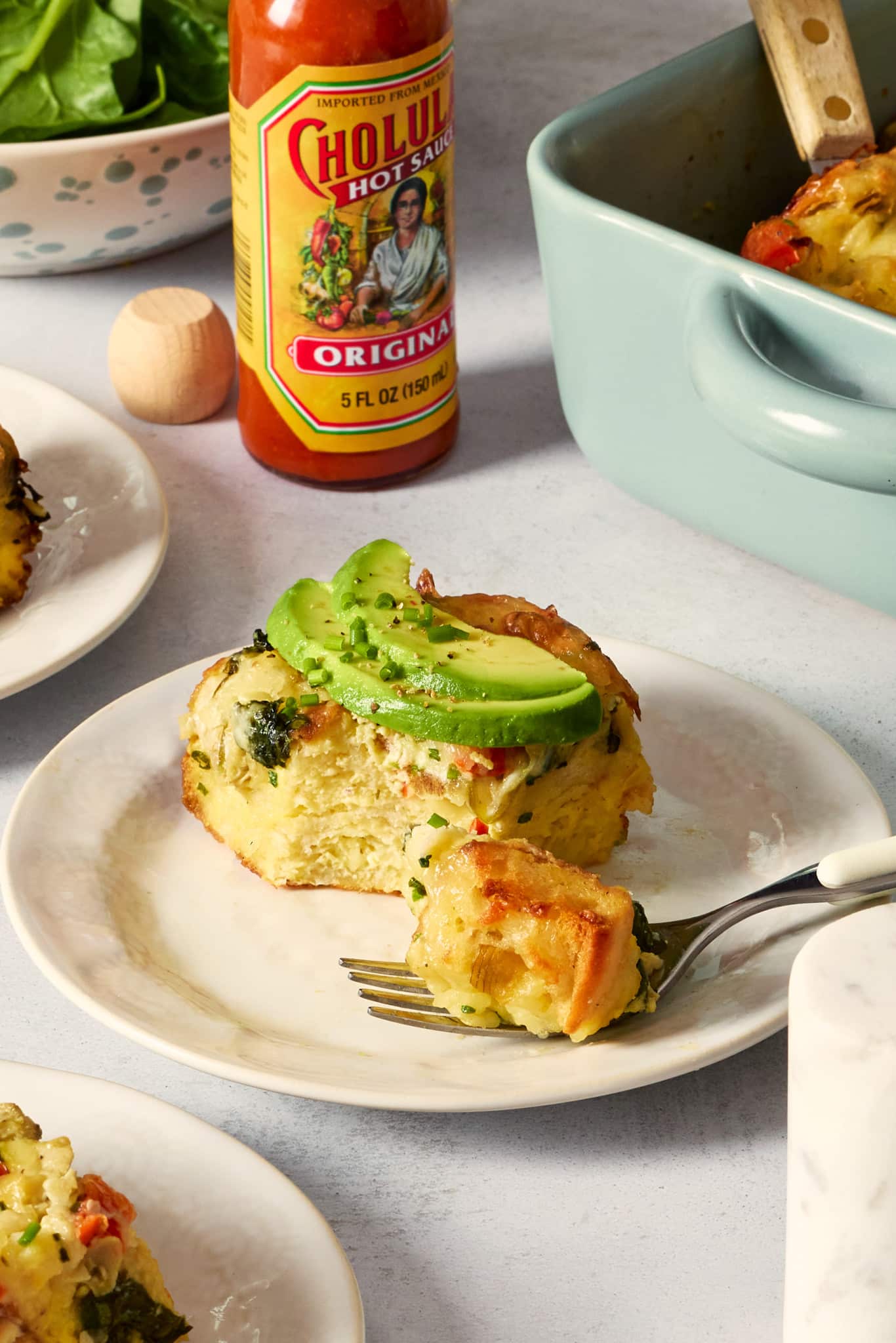 Photo of a plated slice of overnight artichoke spinach egg bake topped with sliced avocado and fresh chives. A fork holding a bite of the casserole sits in front of the plate with a casserole dish and hot sauce bottle in the background.