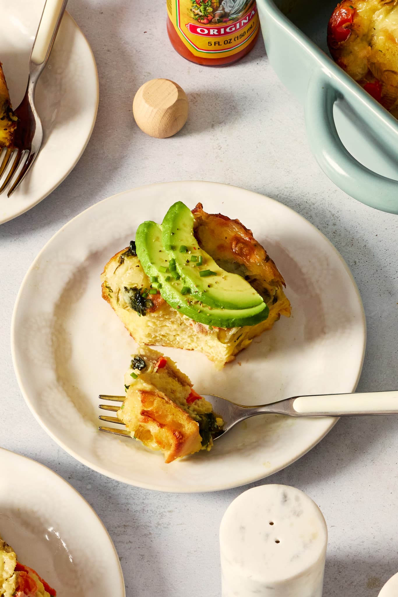 Overhead photo of a plated slice of overnight artichoke spinach egg bake topped with sliced avocado and chives. A fork holding a bite of casserole rests on the plate beside the serving.