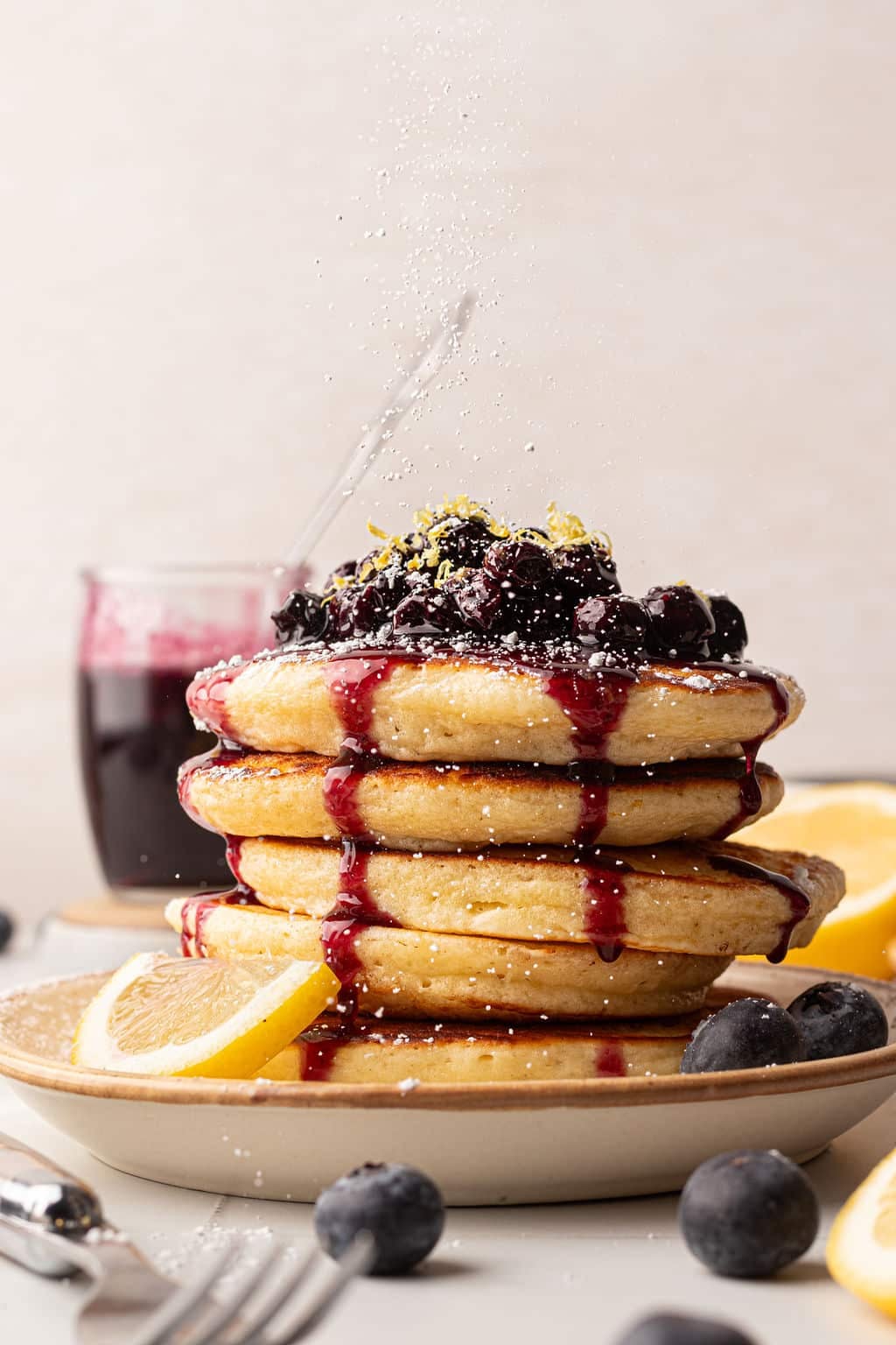 A stack of fluffy pancakes topped with blueberry compote, lemon zest, and blueberry syrup. A light dusting of powdered sugar is falling from above, creating a snowy effect. The plate is surrounded by fresh blueberries and lemon slices.
