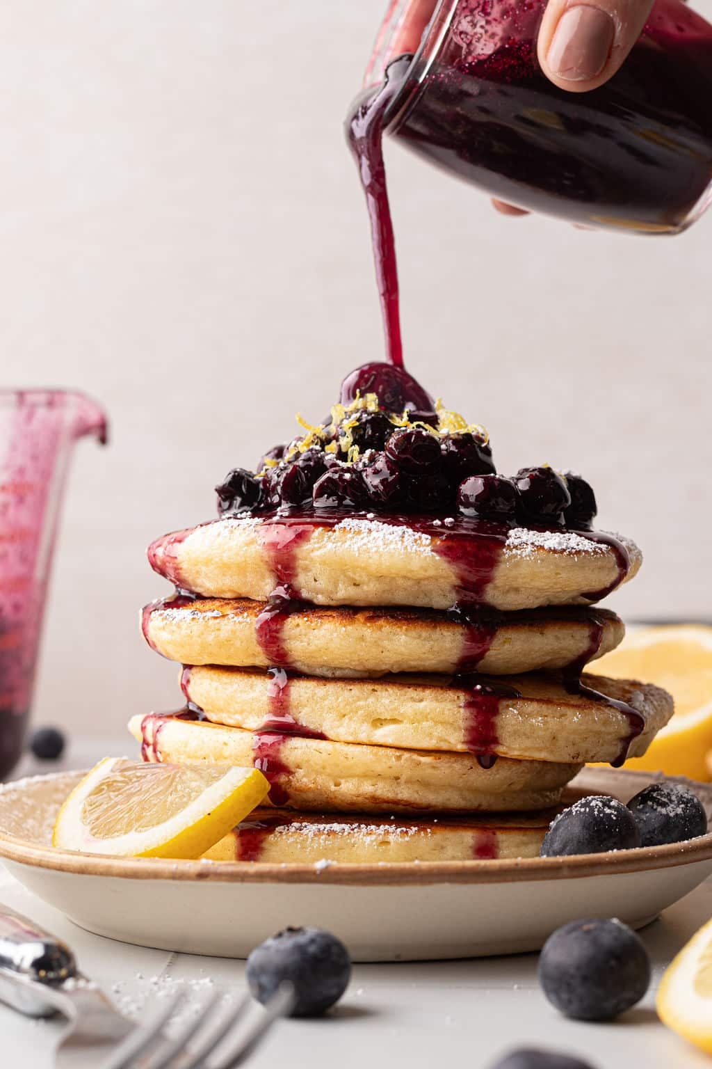 A hand pouring a rich, dark blueberry sauce over a stack of pancakes already topped with blueberries and lemon zest. Powdered sugar is lightly dusted on the pancakes. A jar of sauce and lemon slices are in the background.
