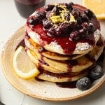 A top-down view of a pancake stack covered in blueberry compote, powdered sugar, and lemon zest. The plate is garnished with fresh blueberries and a lemon wedge. Blueberry sauce has pooled at the base of the pancakes.