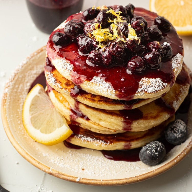 A top-down view of a pancake stack covered in blueberry compote, powdered sugar, and lemon zest. The plate is garnished with fresh blueberries and a lemon wedge. Blueberry sauce has pooled at the base of the pancakes.