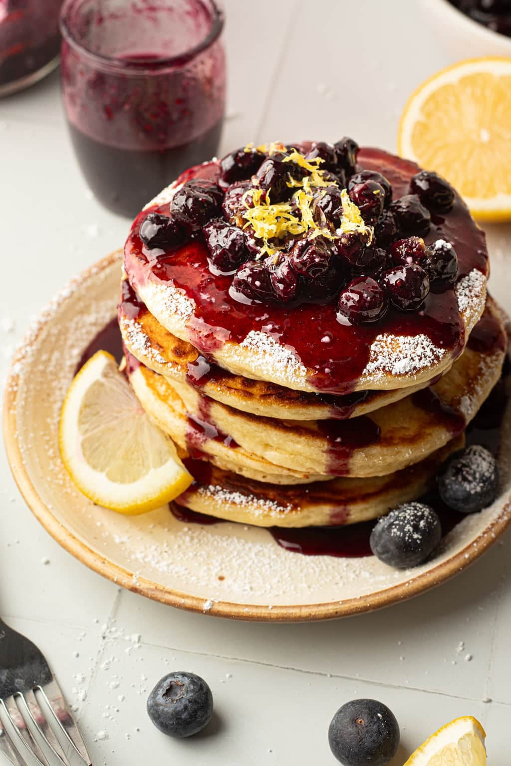 A top-down view of a pancake stack covered in blueberry compote, powdered sugar, and lemon zest. The plate is garnished with fresh blueberries and a lemon wedge. Blueberry sauce has pooled at the base of the pancakes.